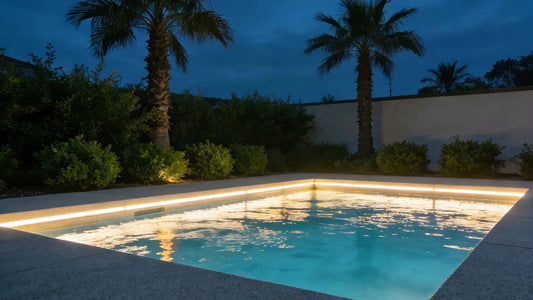 A modern luxury backyard pool at dusk, perfectly illuminated by a warm white LED strip light under the coping, highlighting the water and surrounding palm trees and green hedges.